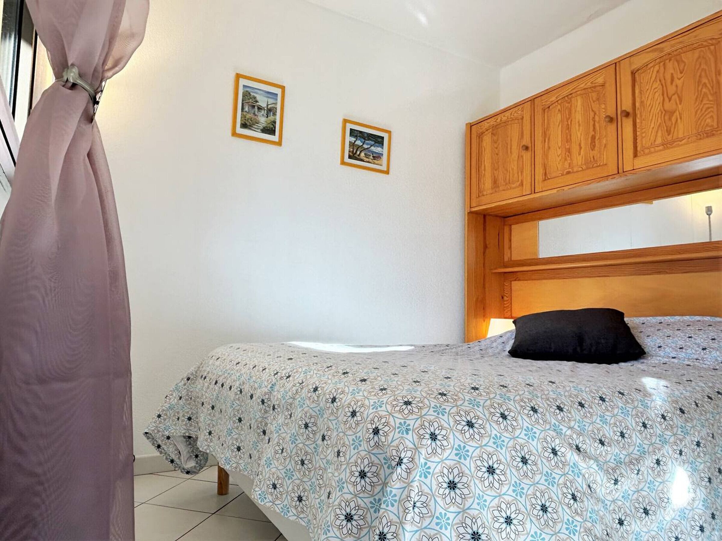 A bedroom with a bed, wooden headboard, two framed pictures on the wall, and a curtain.