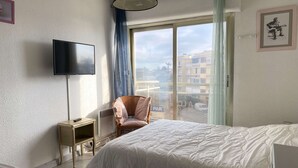 Un dormitorio con cama, televisor, sillón y ventana con vistas a edificios.