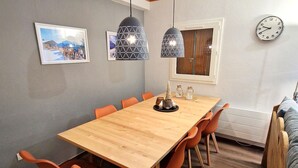 Un coin repas avec une table en bois, des chaises orange, des luminaires suspendus, des tableaux encadrés et une horloge au mur.