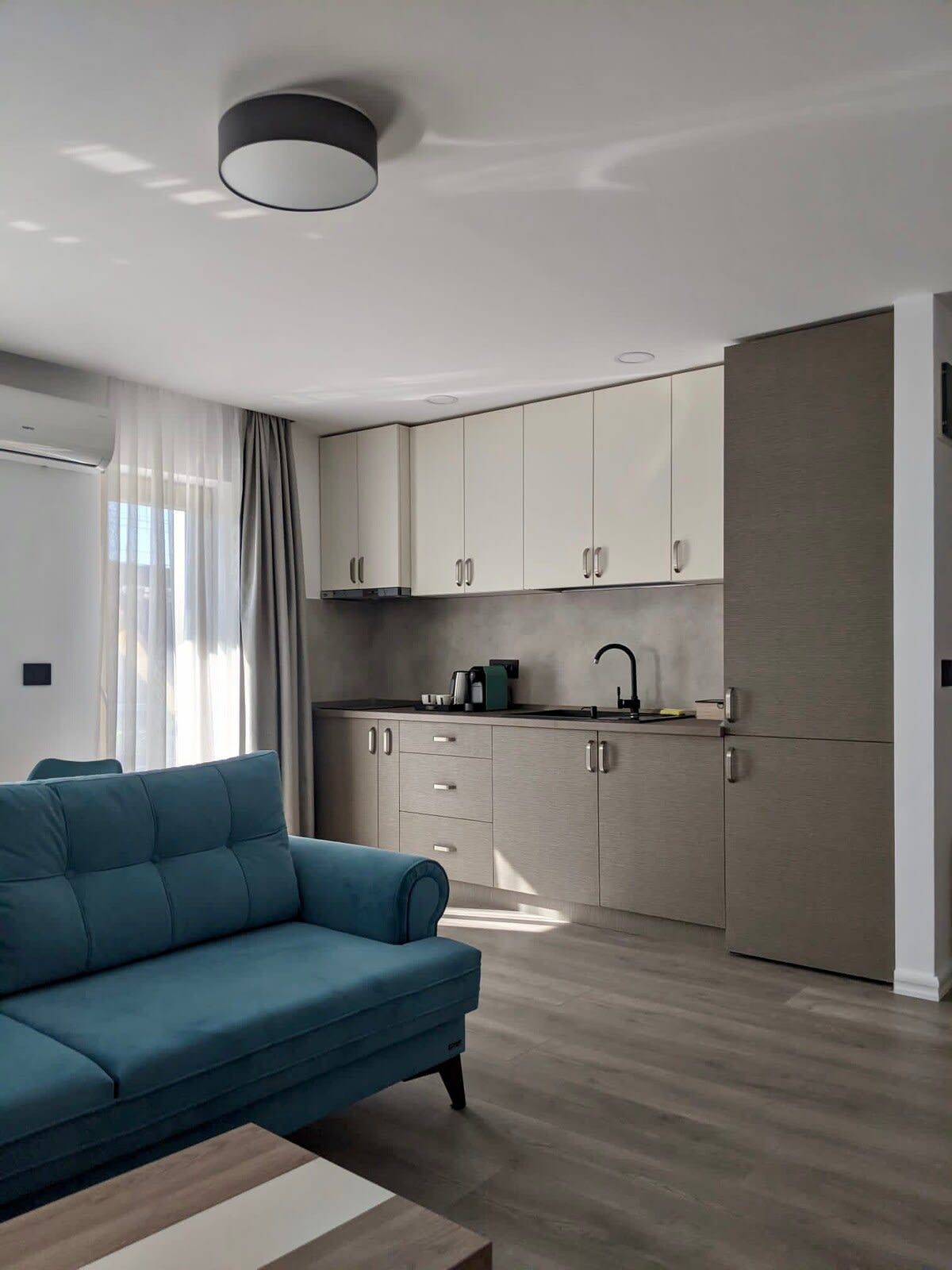 A modern kitchen with light-colored cabinets, a built-in oven, and a sink. There is a blue sofa and a wooden coffee table in the living area.