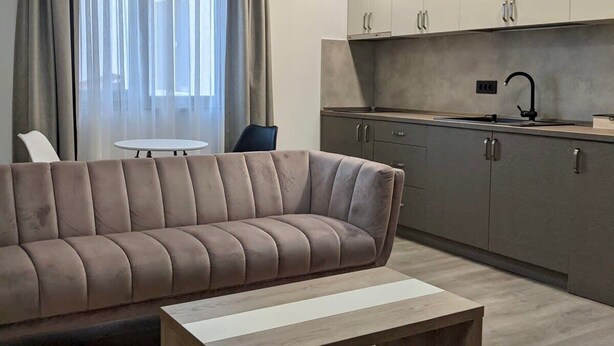 Ein modernes Wohnzimmer mit einem beigefarbenen Sofa, einem Holztisch und einer Küchenzeile mit grauen Schränken und einer Spüle.
