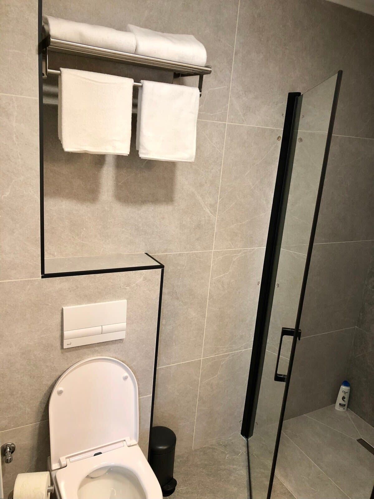 Un baño moderno con inodoro, un perchero con dos toallas, un cesto de basura y una zona de ducha.