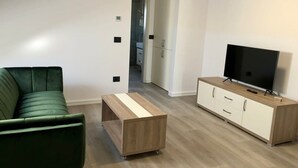 Ein modernes Wohnzimmer mit einem grünen Sofa, einem Holztisch, einem Fernseher auf einer Holzkommode und einem Flur, der zu einem Badezimmer führt.