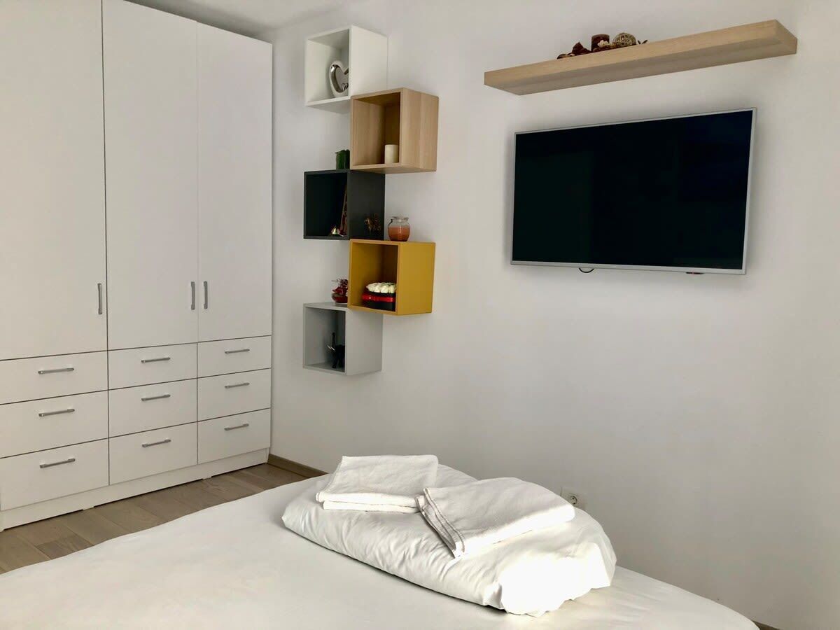 A bedroom with a bed, white wardrobe, shelves with various items, and a TV on the wall.