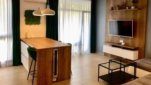 Un soggiorno moderno con zona pranzo in legno, un tavolino da caffè in vetro, una TV a schermo piatto montata su una parete di legno e una pianta verde sulla parete.