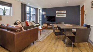 A modern living room with a brown leather sofa, a wooden dining table with chairs, a flat-screen TV mounted on the wall, and a large window with curtains. - Luxury Two Bedroom Apartment Apt 4 (York)