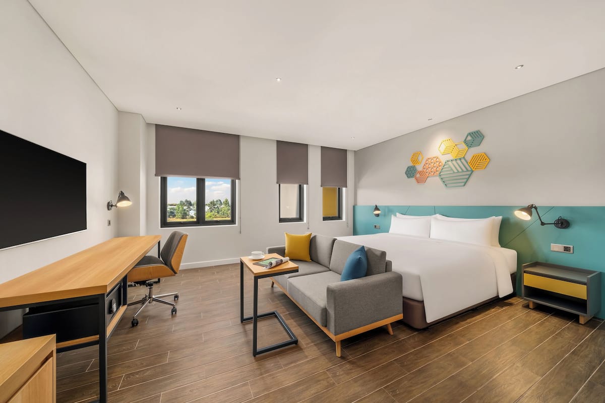 A modern hotel room with a wooden desk, a sofa, a bed, and a nightstand.