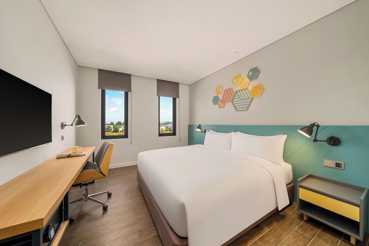 A modern hotel room with a large bed, a desk, and a chair. There are windows providing natural light and a view of the outdoors.