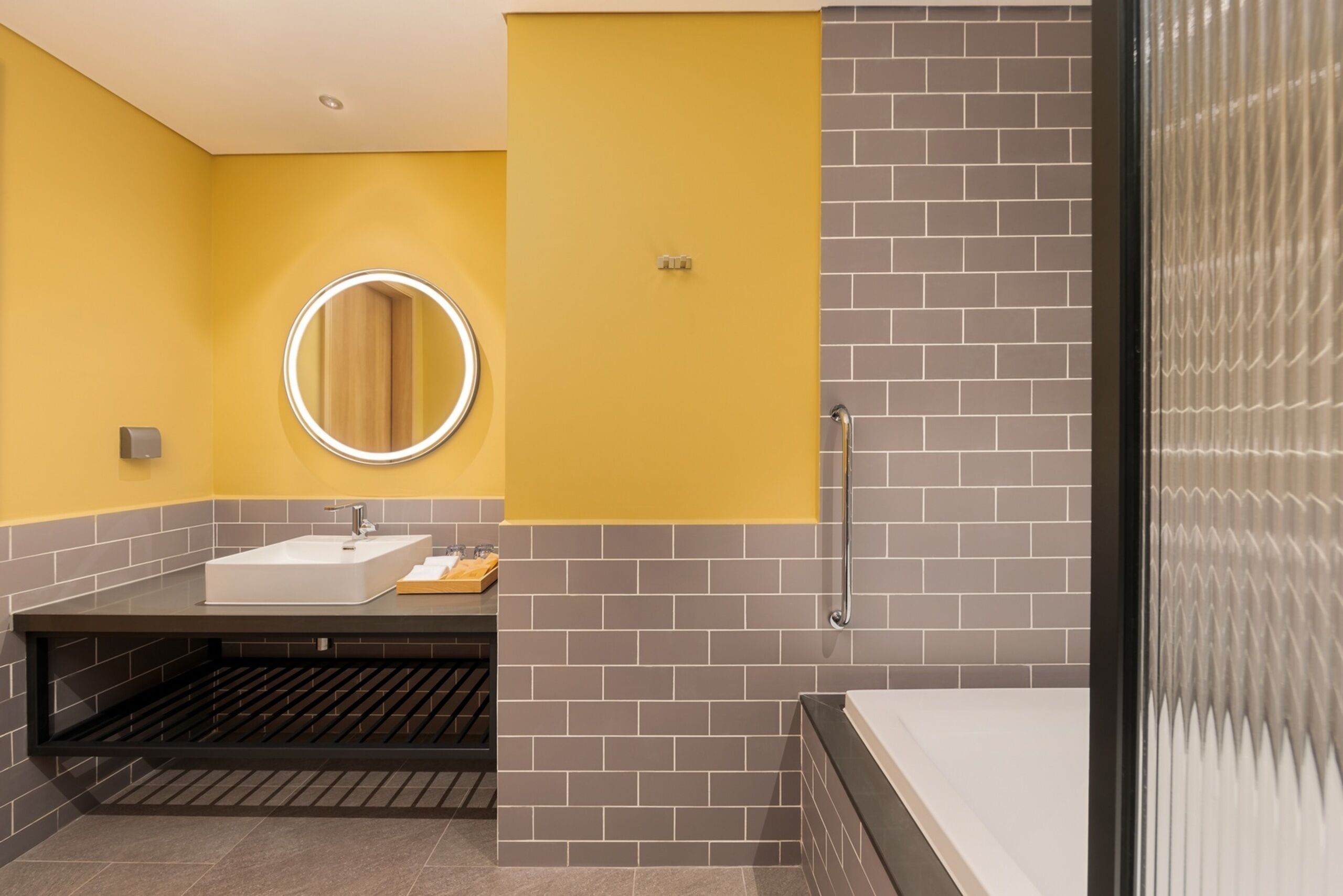 Un baño moderno con una pared de acento amarilla, un espejo redondo con luz, un lavabo blanco y una bañera.