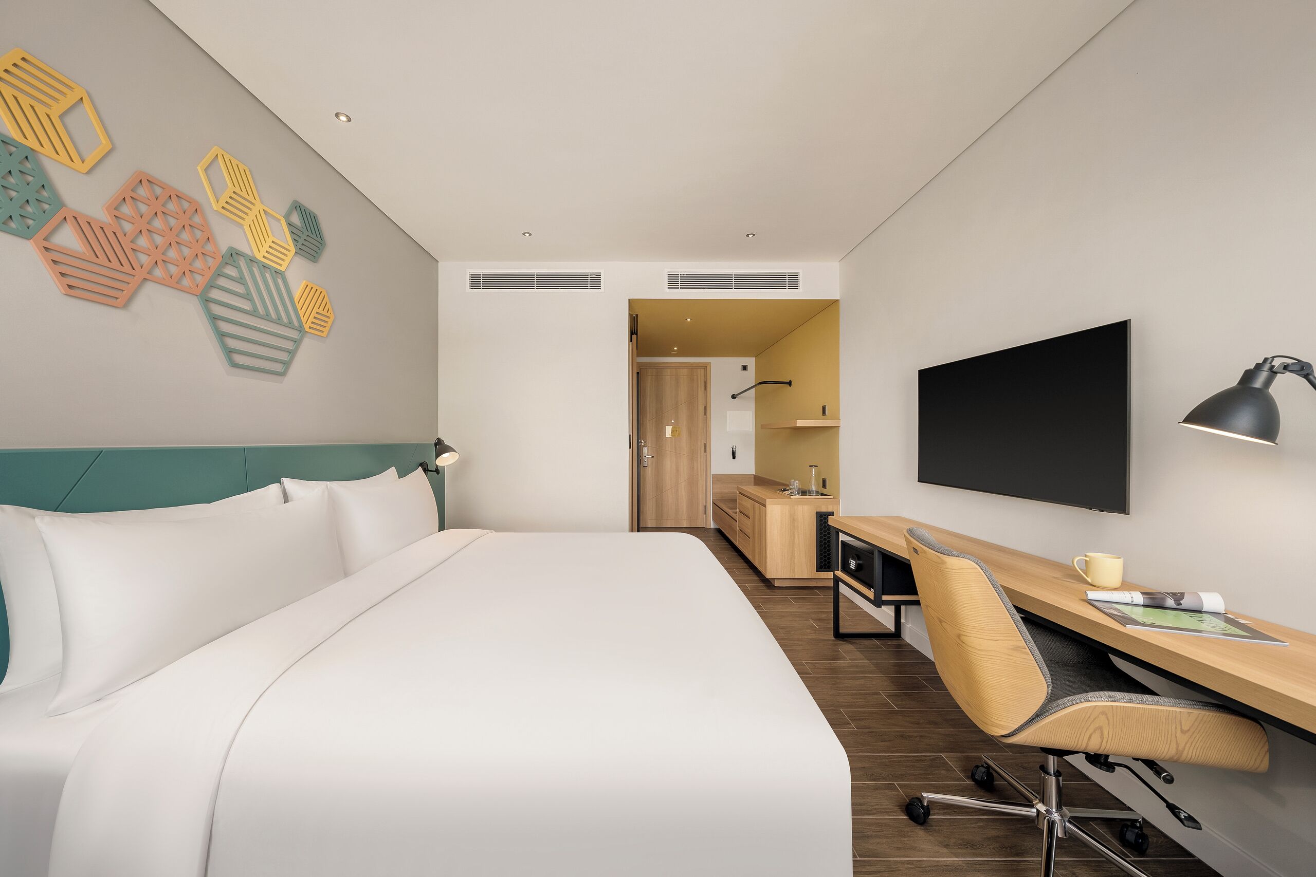 Ett modernt hotellrum med en stor säng, ett skrivbord med en stol, en platt-TV och en väggmonterad lampa.
