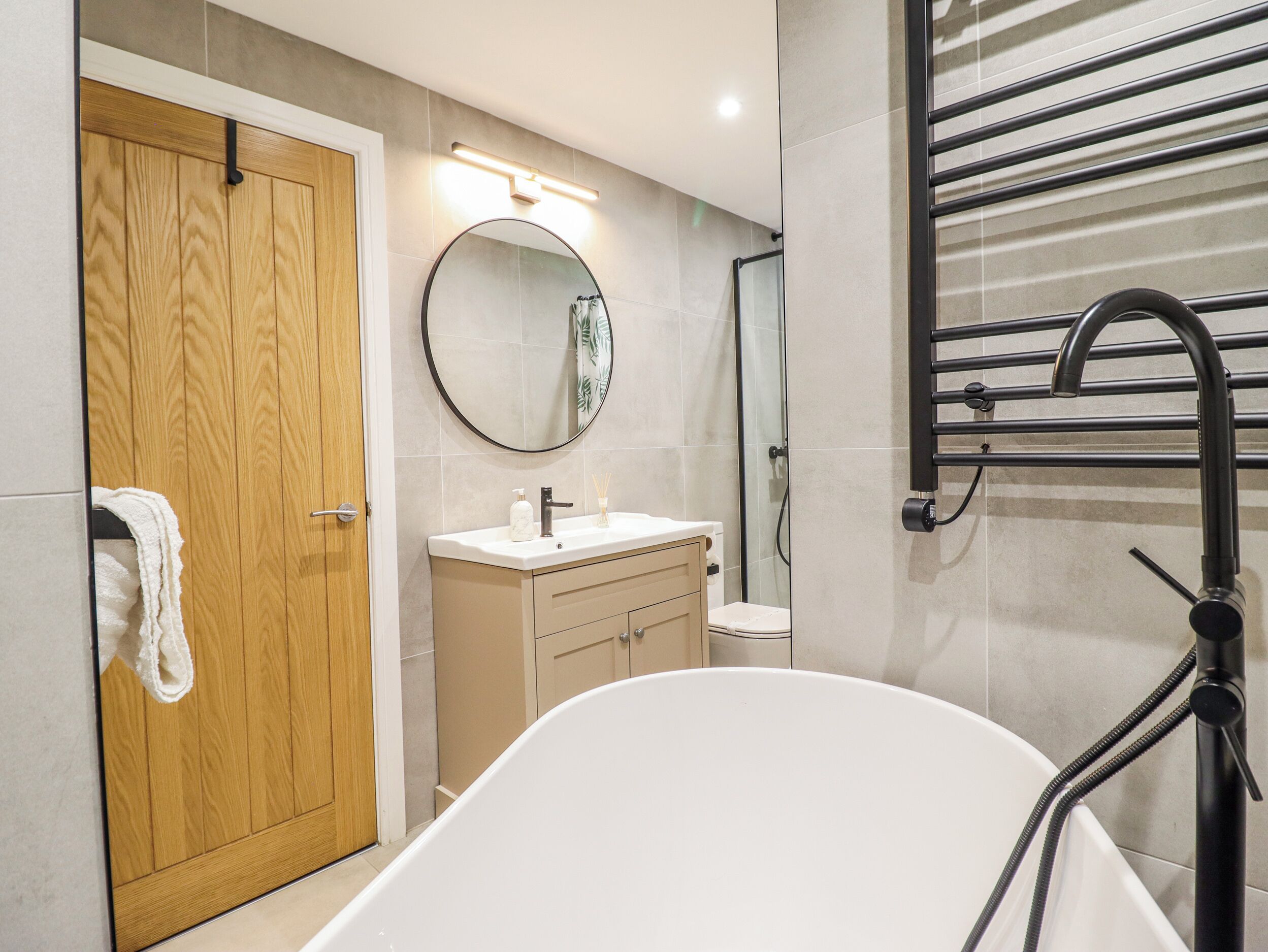 A modern bathroom with a wooden door, a round mirror, a white sink, a bathtub, and a towel rack.
