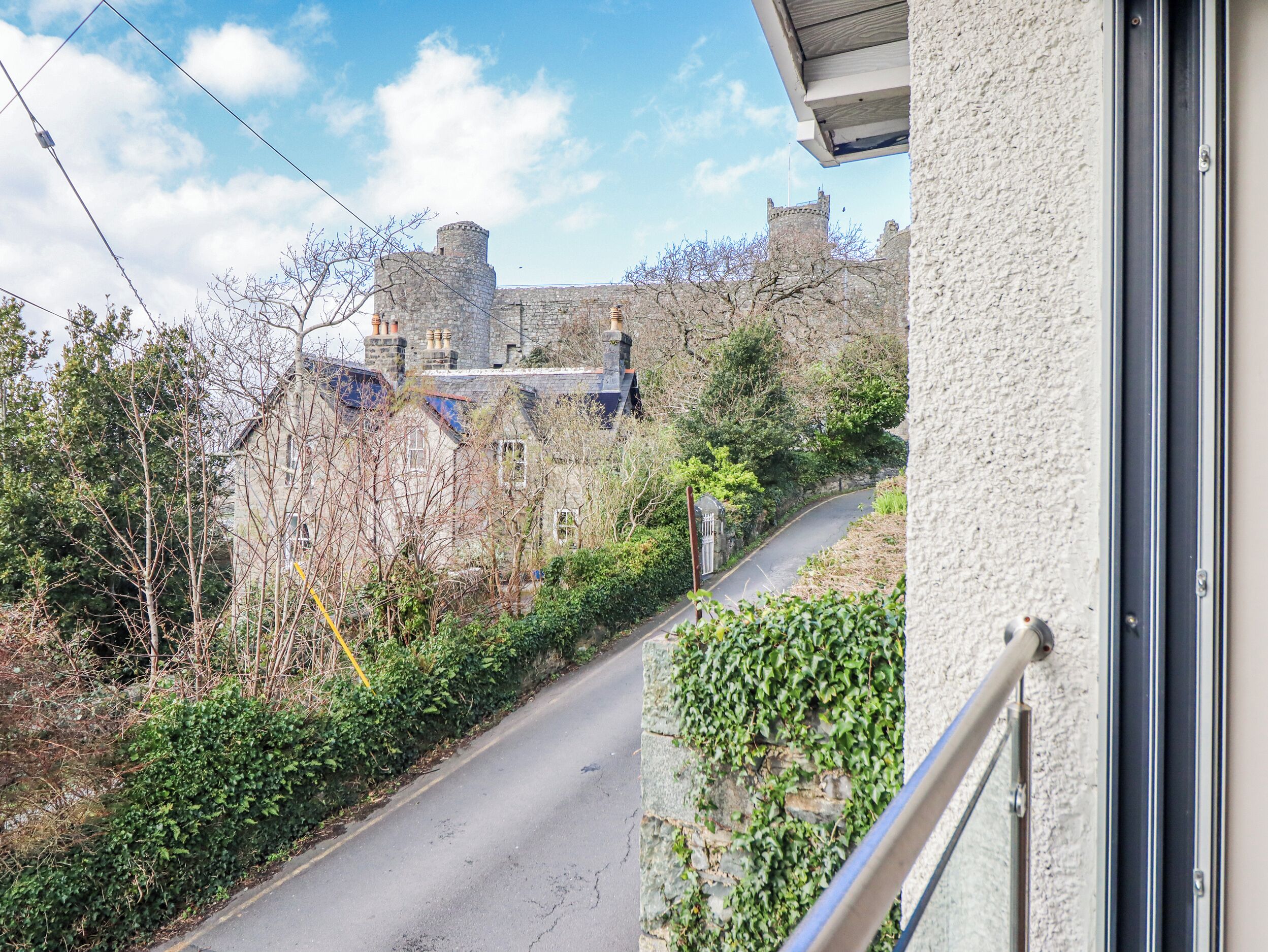A stone building with a turret and a gated entrance, surrounded by greenery and a narrow road.