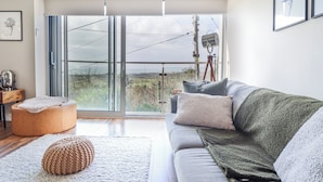 Un salon moderne avec un canapé gris, un pouf en tricot, une table d’appoint en bois et une grande porte-fenêtre coulissante donnant sur un balcon.