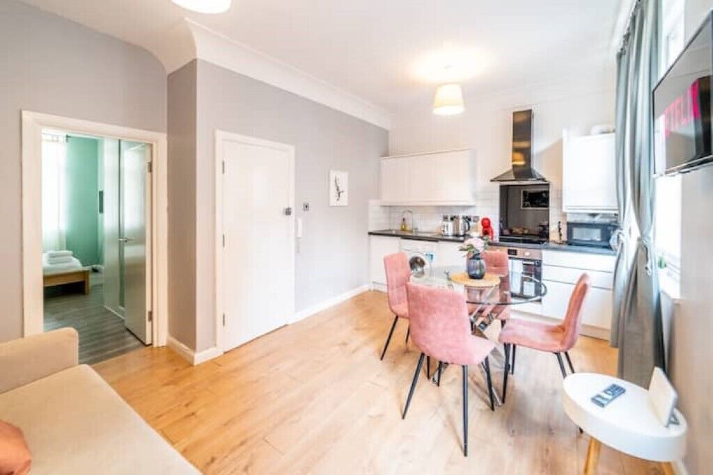 A modern kitchen with white cabinets, a black range hood, and pink chairs around a glass table.