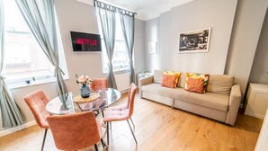 A modern living room with a round glass table, two pink chairs, a beige sofa with colorful cushions, a flat-screen TV, and a framed picture on the wall. - [Covent Garden]Central London Apartment (London)