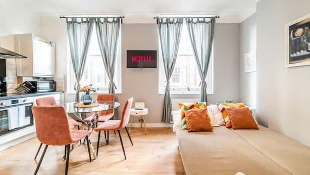 A modern living room with a sofa, a dining table, and chairs. There is a kitchen area with a microwave and cabinets. The room has a TV mounted on the wall and curtains on the windows. - [Covent Garden]Central London Apartment (London)