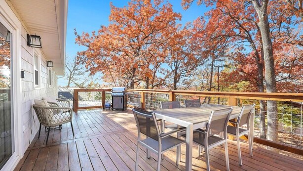 Terraza de madera con mesa y sillas, con vistas a árboles de follaje otoñal.