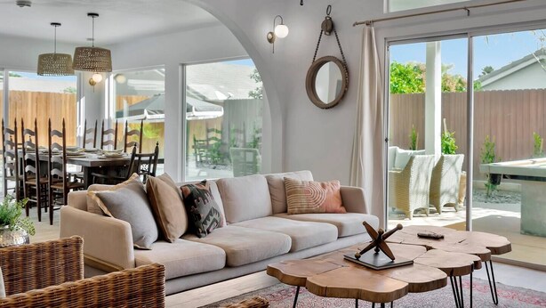 A modern living room with a large sofa, a coffee table, and wicker furniture. There is a dining area with a table and chairs visible through the glass doors. - Desert Rose - Tiki Game-room, 10 min to Indian wells tennis garden Festivals, Golf and more! (Indio)