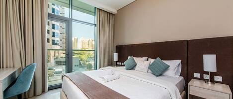 A modern hotel room with a large bed, a desk, and a view of the city through floor-to-ceiling windows.