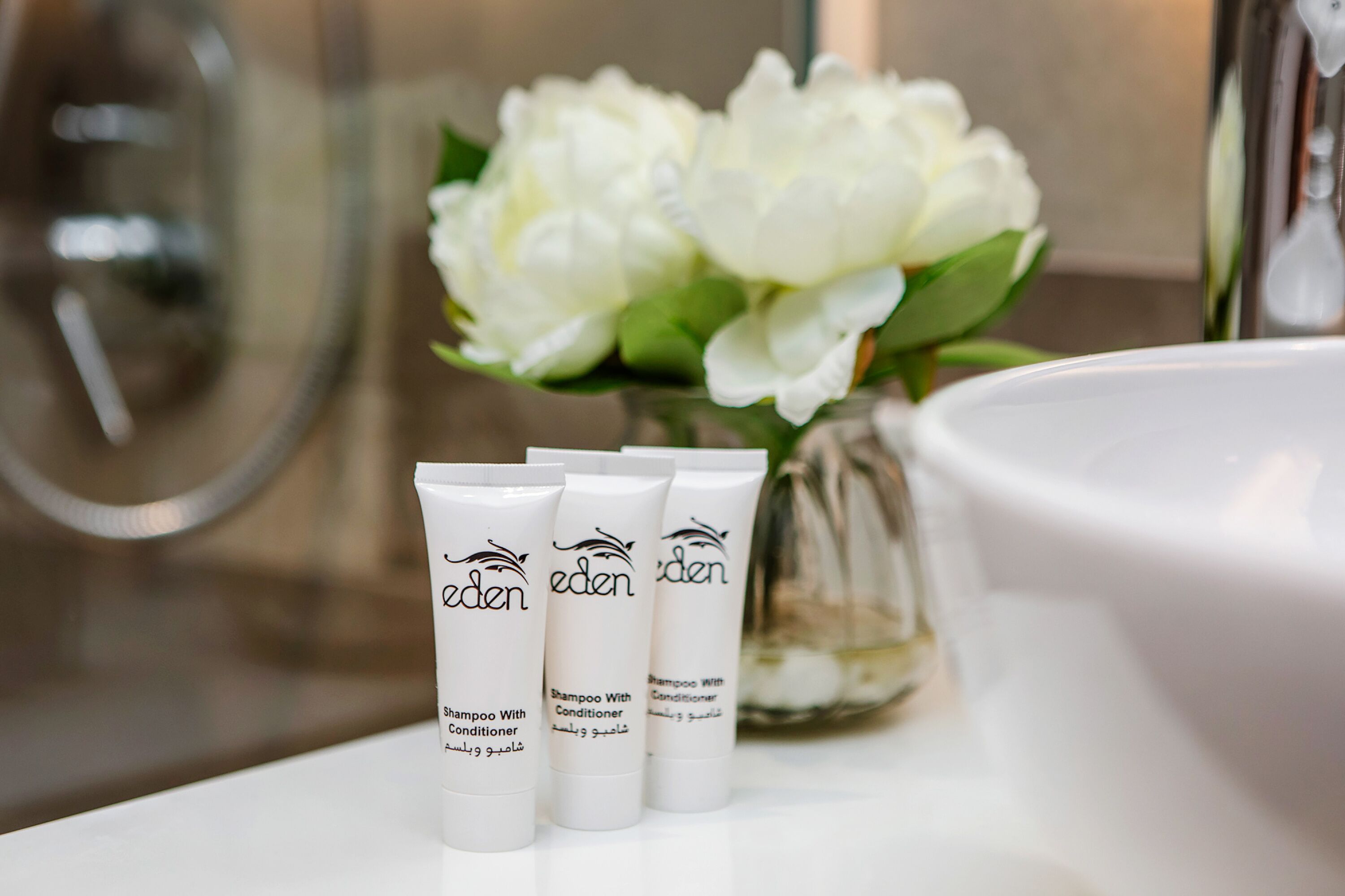 Drie flessen Eden shampoo en conditioner op een wit oppervlak, met een vaas witte bloemen op de achtergrond.