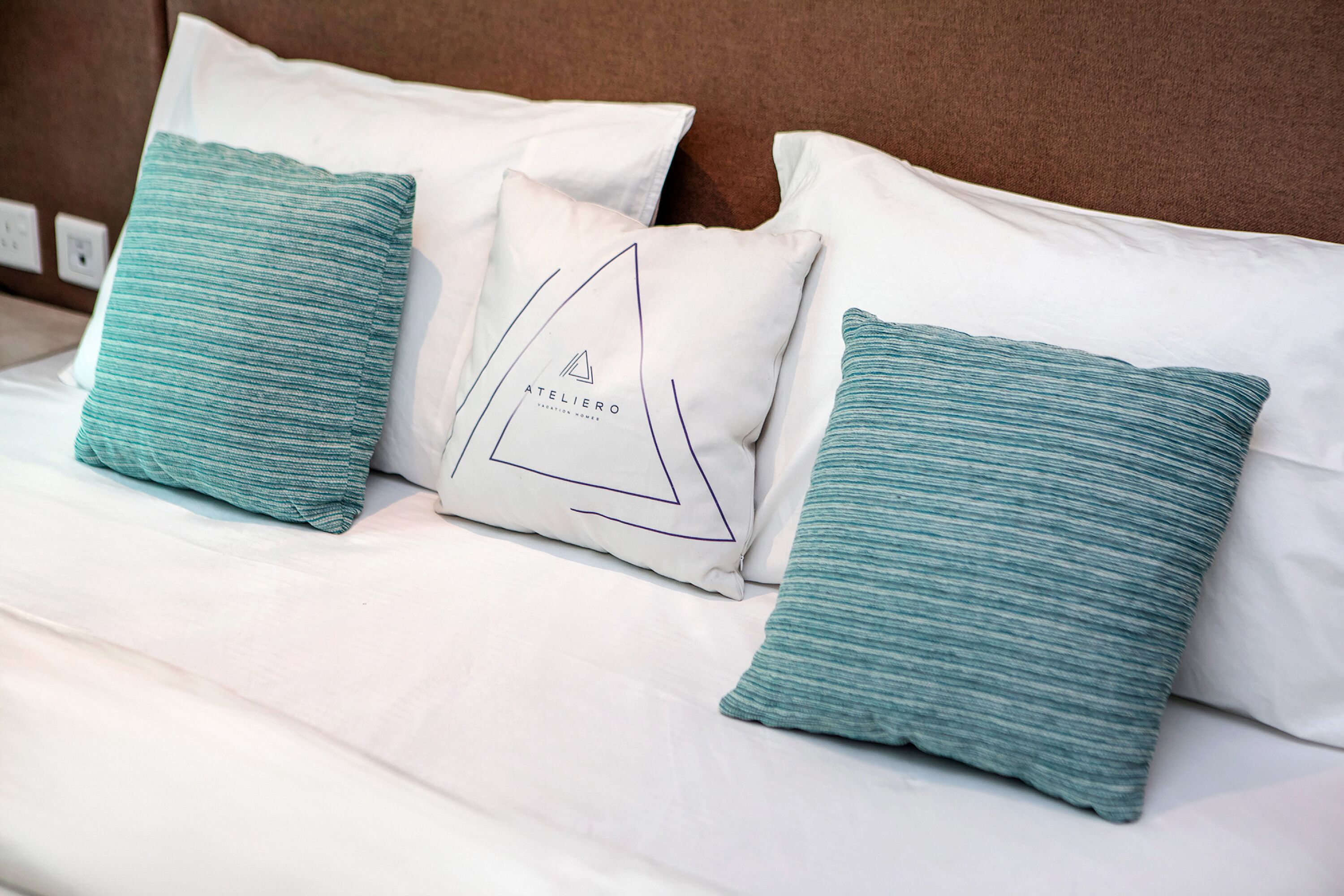A neatly made bed with white linens and three teal pillows, one with a geometric design.