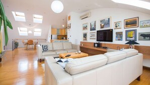 Un salon moderne avec un canapé, une table basse et un téléviseur. Au fond, on aperçoit une cuisine avec un coin repas.