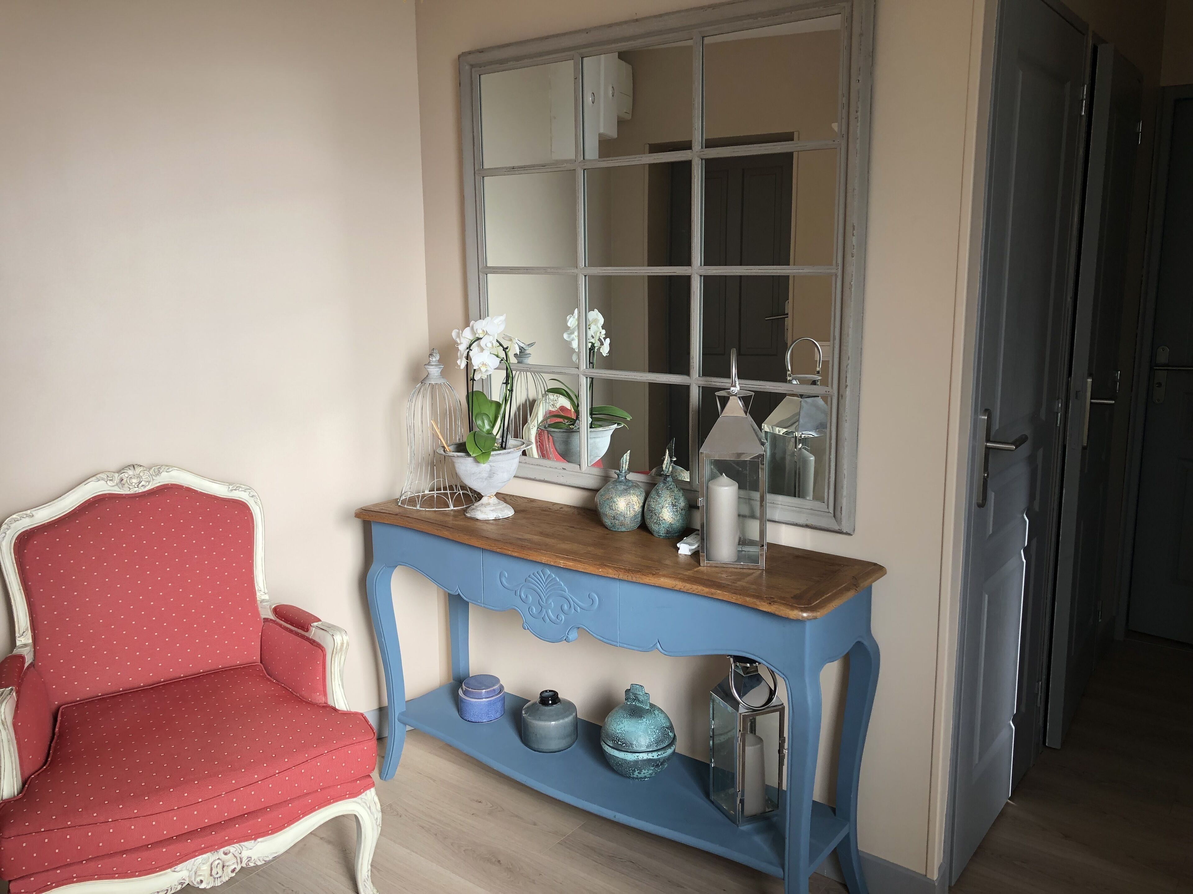 Una habitación con una silla de lunares rojos, una consola azul con objetos decorativos y un espejo encima.