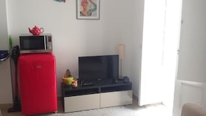 Un saló amb un televisor de pantalla plana, un refrigerador vermell, un sofà beix i una taula petita amb diversos objectes.