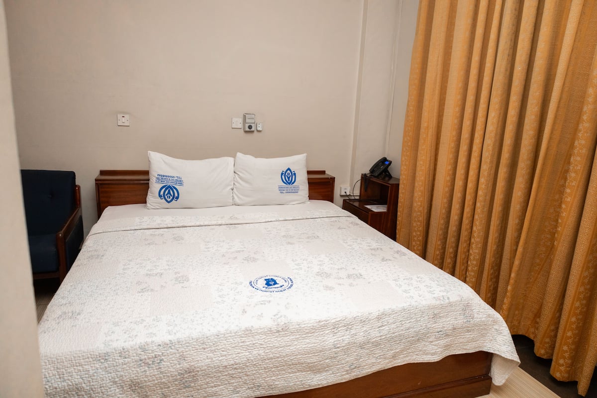 Habitación de hotel con cama, dos almohadas, mesita de noche, teléfono y cortina.