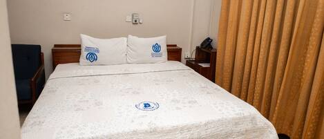 A hotel room with a bed, two pillows, a bedside table, a phone, and a curtain.