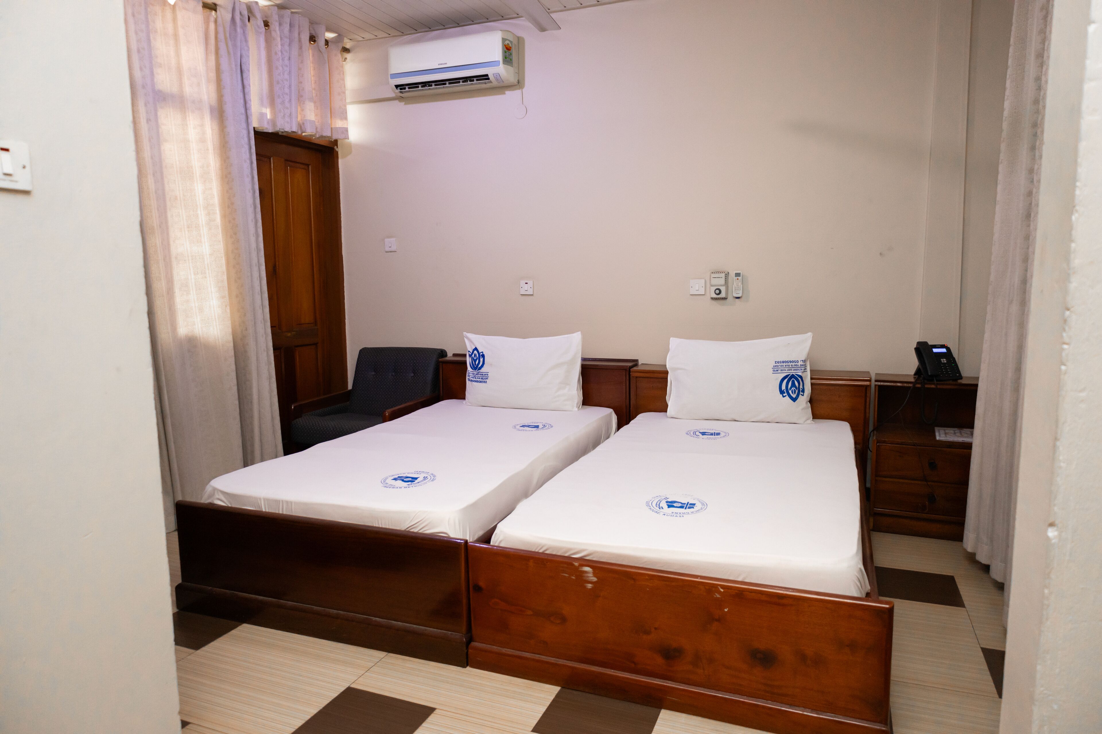 A room with two single beds, a wooden wardrobe, a desk with a chair, and an air conditioning unit on the wall.