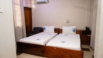 A room with two single beds, a wooden wardrobe, a desk with a chair, and an air conditioning unit on the wall.