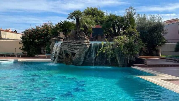 Una piscina con caída de agua rodeada de abundante vegetación y un edificio al fondo.