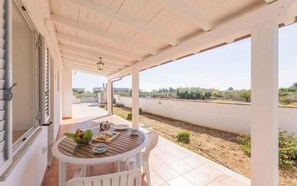 Une terrasse extérieure couverte, avec une table dressée pour deux personnes, donnant sur un espace paysager aménagé et offrant une vue dégagée sur les bâtiments situés au loin.