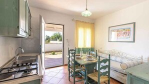 Una cocina con estufa, lavamanos y mesa de comedor con sillas. Hay una ventana con cortinas, una cama con colcha estampada, y un cuadro en la pared.