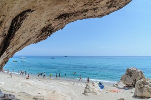 A beach with a large rock formation arching over the water, people walking along the shore, and boats in the distance.