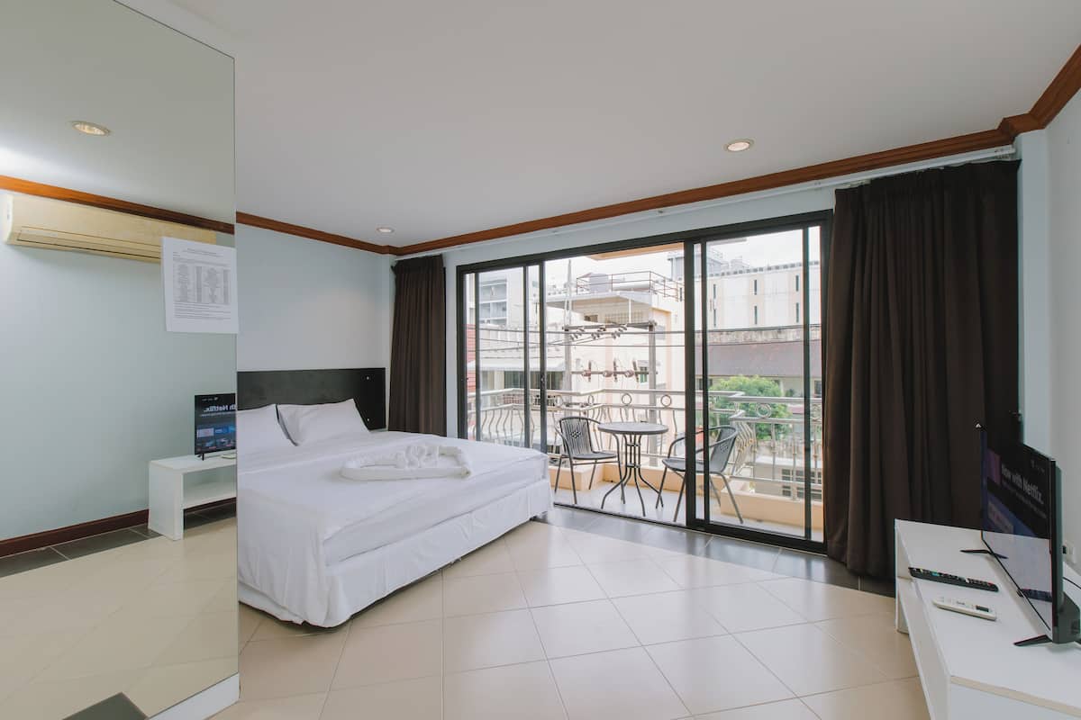A hotel room with a bed, a desk with a TV, a balcony with chairs, and a view of buildings.