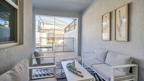 A modern living room with a sofa, armchairs, and a coffee table. There is a large window providing a view of a balcony with outdoor furniture.