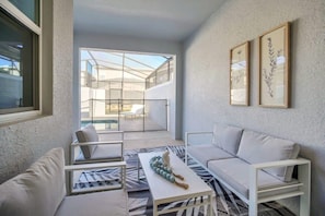 A modern living room with a sofa, armchairs, and a coffee table. There is a large window providing a view of a balcony with outdoor furniture.