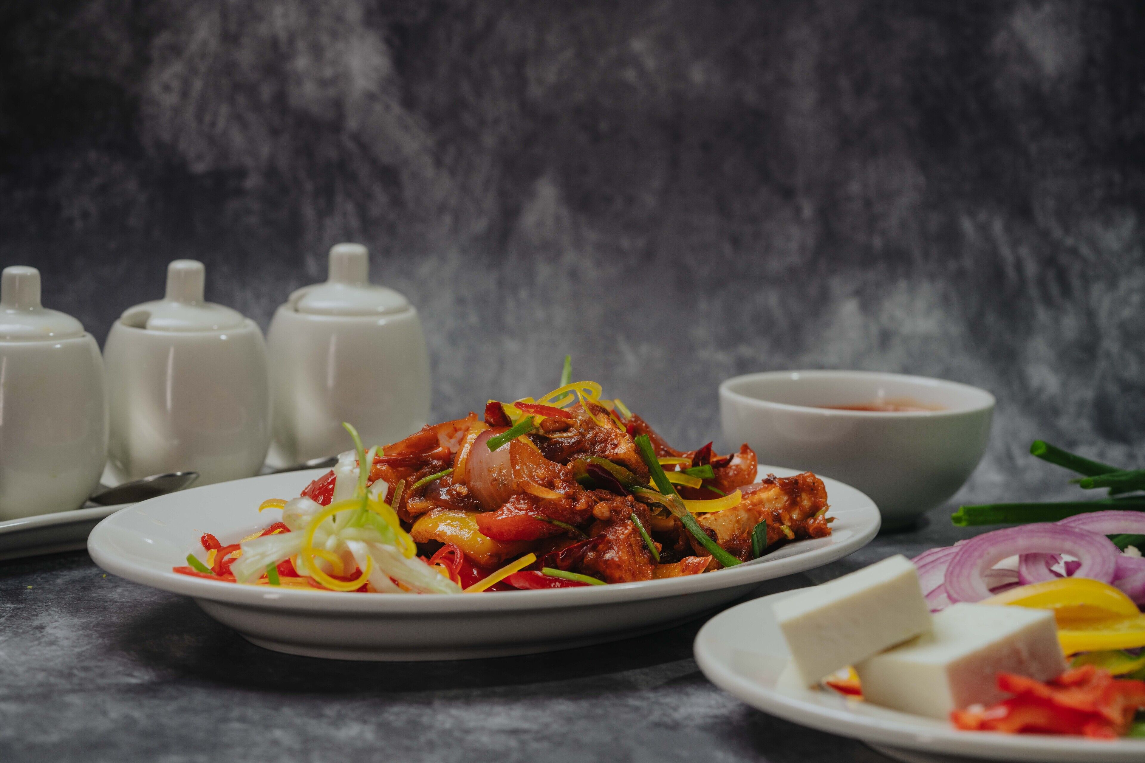 A plate of food with various vegetables and meat, accompanied by a bowl of sauce and a plate of cheese.