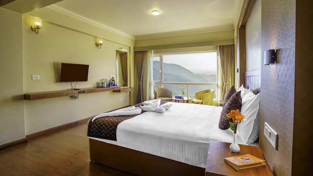 A hotel room with a large bed, a television on the wall, a window with a view of mountains, and a bedside table with a book and a vase of flowers.