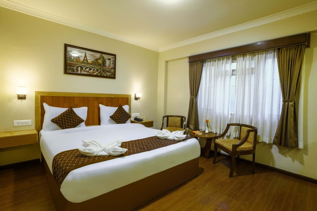 A hotel room with a large bed, a wooden headboard, a nightstand, a chair, a small table, and a painting of a cityscape.