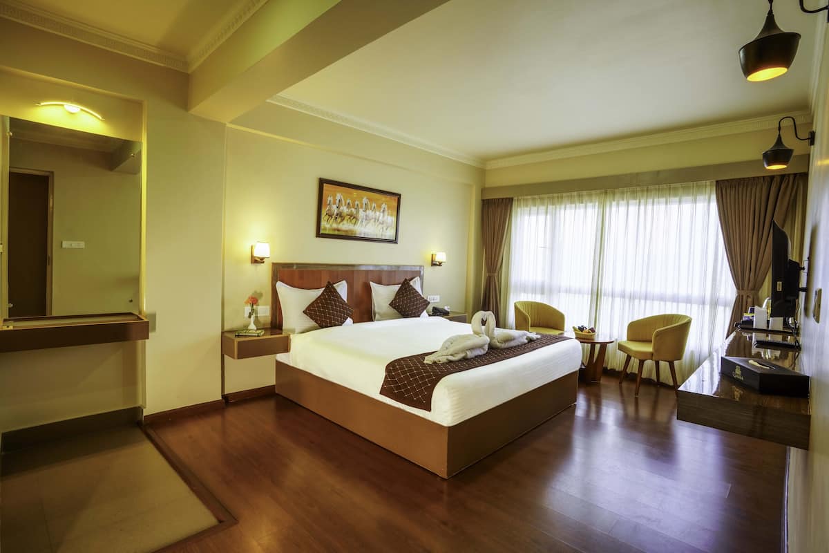 A hotel room with a large bed, a wooden floor, a desk with a chair, a small table with a vase, and a painting on the wall.