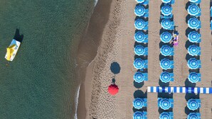 Une vue aérienne d’une plage avec des rangées de parasols bleus, un parasol rouge et un bateau.