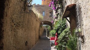 A narrow stone alleyway with a rustic stone wall on one side and a building with a wooden balcony on the other. - Mobilehome with air conditioning, pool in a sunny location (Castiglione della Pescaia)