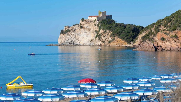 A coastal area with a castle on a cliff, a boat, and beach umbrellas. - Mobilehome with air conditioning, pool in a sunny location (Castiglione della Pescaia)