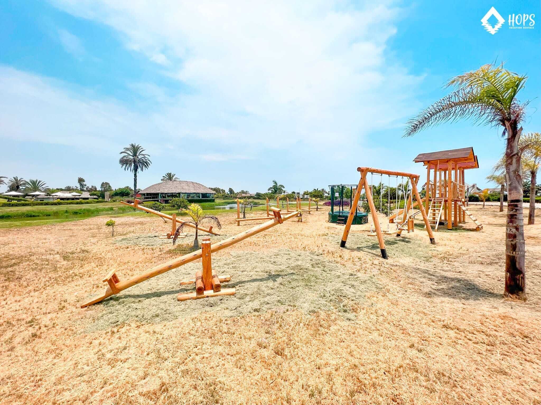 Un parque infantil con columpios y una estructura para escalar en una zona de arena, palmeras y un edificio al fondo.