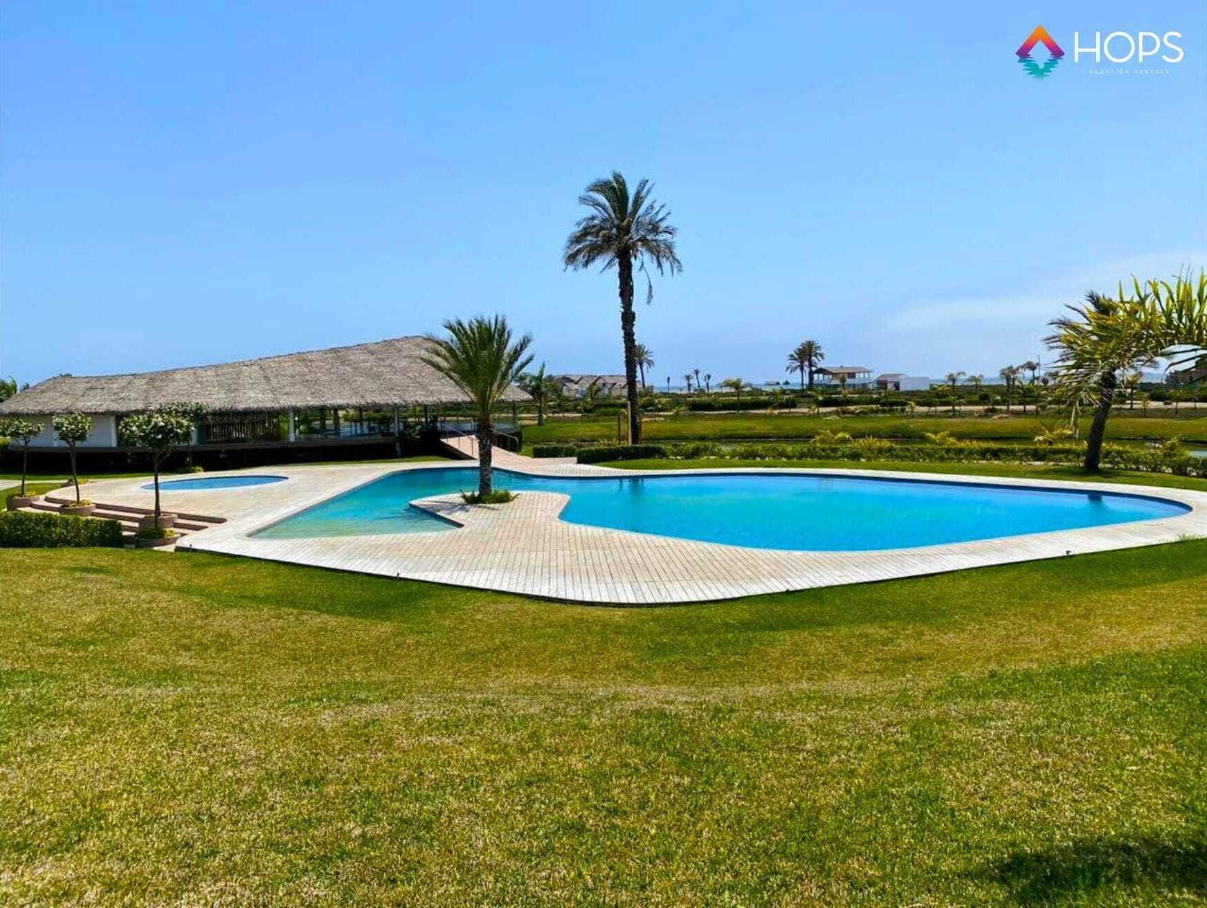 Une piscine dotée d’une terrasse en bois, entourée de pelouse et de palmiers, avec un bâtiment visible au loin.