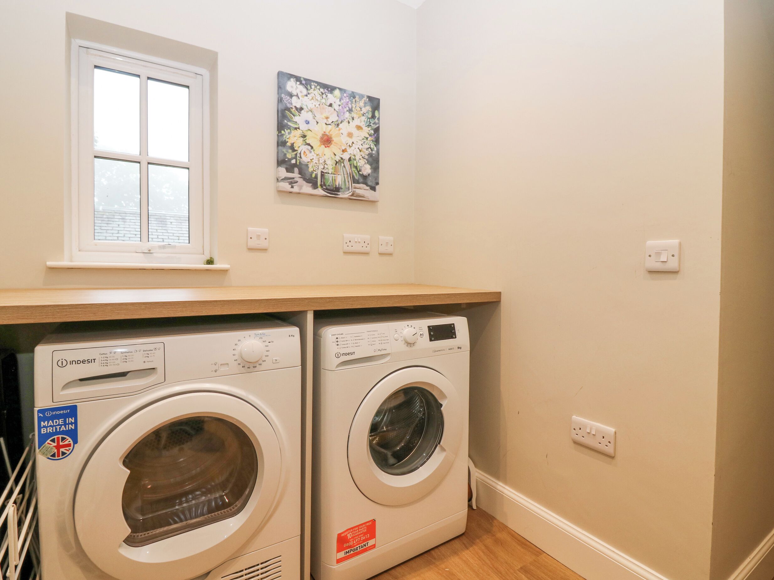 A laundry room with two white Indesit washing machines, a window, and a floral painting on the wall.