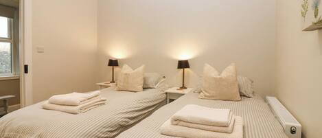 Habitación de hotel con dos camas, cada una con ropa de cama y almohadas a rayas, una mesita de noche con lámpara y un radiador.